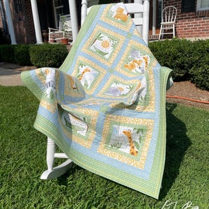 May include: A handmade baby quilt with a safari animal theme. The quilt features squares with giraffes, lions, and other animals in a blue, yellow, and green color scheme. The quilt is draped over a white rocking chair on a grassy lawn.