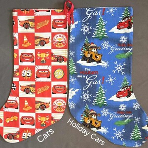 May include: Two Christmas stockings, one red with a Cars theme and the other blue with a Cars holiday theme. The red stocking has a checkered pattern with red race cars and the text "95", "GO", "CHAMP", and "ACCELERATE". The blue stocking has a snowflake pattern with the text "Gas!" and "The Holidays are a Gas!"
