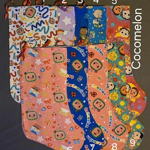 May include: Nine colorful Christmas stockings with various Cocomelon-themed patterns. Designs include musical notes, rainbows, characters, and watermelon graphics. Each stocking has a loop for hanging and is made of fabric.