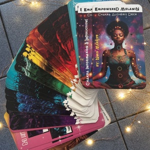 May include: A deck of cards with a colorful rainbow design. The cards are fanned out in a semi-circle. The top card features a woman meditating with a dark background and the text "EMPOWERED MELANIN CHAKRA ALCHEMY DECK BY: NIKO MERRITT".