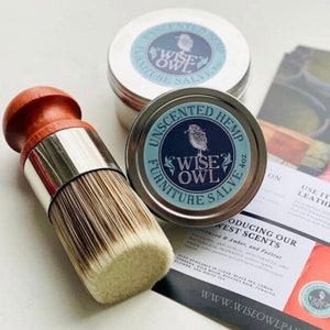 May include: Two tins of Wise Owl Furniture Salve, one unscented hemp and one with a West Scents fragrance. The tins are silver with blue and white labels featuring an owl illustration. A wooden-handled brush with natural bristles is also pictured. The text on the label reads "Wise Owl Furniture Salve 4 oz."