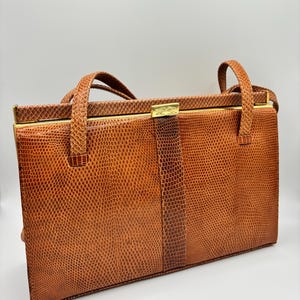 Może przedstawiać: Vintage brązowa torebka o fakturze skóry gada. Prostokątna torba posiada złoty zatrzask i uchwyty. Torba ma około 30 cm szerokości i 20 cm wysokości.