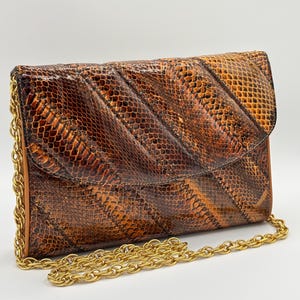 May include: A brown and orange snakeskin-patterned clutch bag with a gold chain strap. The flap closure has a diagonal quilted design. The bag is rectangular and appears to be made of leather.