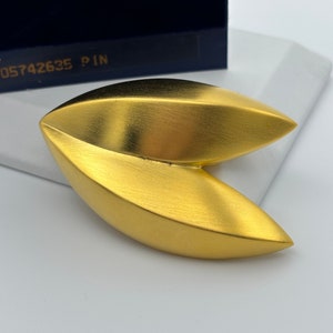 May include: A gold-colored metal pin shaped like a fortune cookie. The pin is on a white surface.