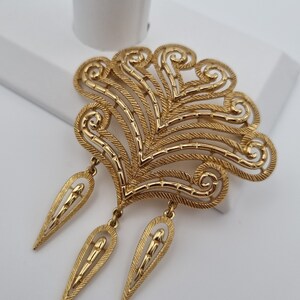 May include: A gold-toned brooch with an ornate, fan-like design. The brooch features intricate detailing, including rope-like textures and dangling teardrop-shaped pendants. The overall design is symmetrical and elegant, suitable for formal wear.