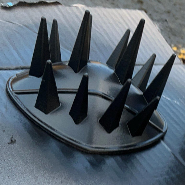 May include: A black, rounded, dome-shaped accessory featuring a series of twelve pointed, cone-like spikes protruding from the top surface. The object has a smooth, matte finish and a defined, raised border around the base perimeter.