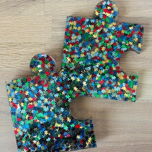 May include: Two clear resin jigsaw pieces filled with colourful glitter. The glitter is in the shape of small jigsaw pieces and is blue, green, red, and gold.