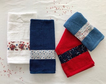 May include: Three kitchen towels with red, white, and blue patriotic designs. The towels are folded and arranged on a white surface.