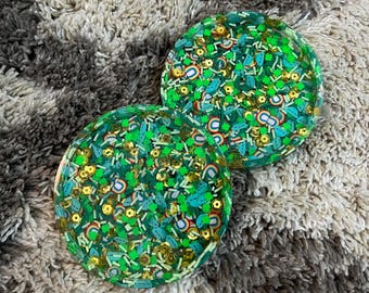 May include: Two round green and gold coasters with a variety of green, gold, and white sprinkles. The coasters are on a gray and white fuzzy surface.