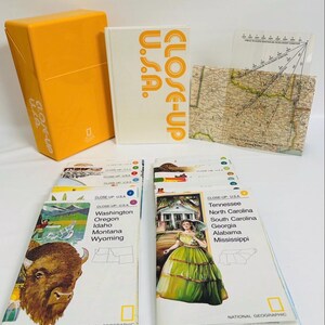 May include: A National Geographic Close-Up U.S.A. map set featuring a yellow storage box, a white hardcover book, a transparent plastic map overlay, and multiple folded paper maps with illustrations of a bison and a person in a green dress.