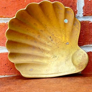 May include: A vintage brass seashell-shaped dish with a scalloped edge. The dish has a golden hue with signs of age and wear. The background features a red brick wall and a brown surface.