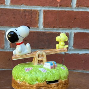 May include: A ceramic music box featuring Snoopy and Woodstock on a seesaw. Snoopy is white with a black ear and Woodstock is yellow. The base is green with flowers and a small gift. A pink balloon is attached.