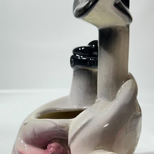 May include: A ceramic vase in the shape of a seated figure, with a black and white color scheme. The figure's hands cradle a central opening, with pink accents on the base and interior. The vase has a glossy finish.
