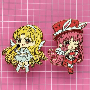 以下が含まれることがあります： アニメスタイルのキャラクターをフィーチャーしたエナメルピン2つ。最初のピンは、マイクと天使の羽根をつけたブロンドの女の子で、青と白のドレスを着ています。2つ目のピンは、ウサギの耳と赤と白のドレスを着たピンク髪の女の子で、星が付いた赤い帽子をかぶっています。