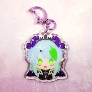 Op de afbeelding: Een sleutelhanger van transparant acryl met een paars en groen anime-personage met een kroon en vleugels. Het personage heeft groen haar en groene ogen en draagt een zwart-witte outfit. De sleutelhanger heeft een zilveren metalen sluiting.