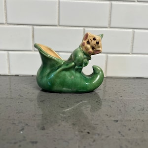 May include: A green ceramic elf shoe planter with a smiling elf face. The elf is peeking out of the shoe.