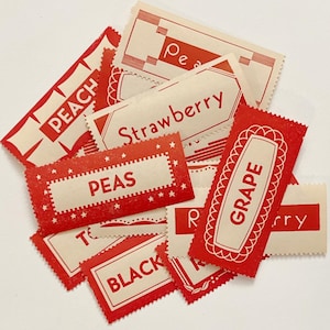 Op de afbeelding: Een verzameling vintage rode en witte voedseletiketten. De labels bevatten tekst zoals "PEACH", "STRAWBERRY", "PEAS", "GRAPE" en "BLACK". De labels hebben decoratieve randen en zijn in een verspreide stapel gerangschikt.