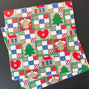 May include: Two square Christmas-themed napkins with a patchwork design. The pattern includes red hearts, green Christmas trees, houses, and angels. The squares are bordered with green and white, and the background is off-white.
