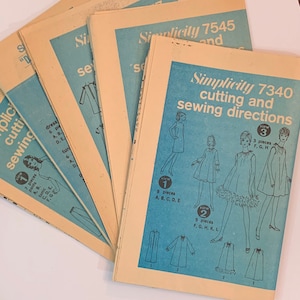 Puede incluir: Una pila de instrucciones de patrones de costura vintage Simplicity para vestidos. El patrón superior es Simplicity 7340, con un fondo azul y dibujos de línea negra de vestidos. El texto "cutting and sewing directions" está impreso en negro.
