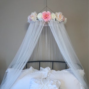 May include: A white canopy bed with a floral crown and sheer white fabric. The bed is made with white bedding and a decorative pillow. The floral crown features pink and white flowers.