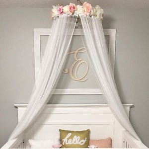 May include: A white crib with a canopy featuring a floral crown and flowing white fabric. The crib is decorated with pink bedding and a gold pillow that says "hello". A wooden letter "E" is on the wall.