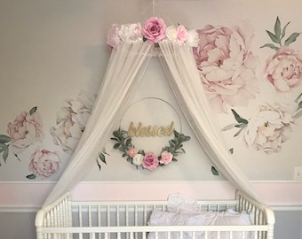 May include: A white crib with a pink ruffled skirt and bedding, set against a wall with large peony decals. A canopy with pink and white flowers and the word "blessed" adds a decorative touch to this nursery.