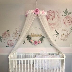 May include: A white crib with a pink ruffled skirt and bedding, set against a wall with large peony decals. A canopy with pink and white flowers and the word "blessed" adds a decorative touch to this nursery.