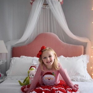 May include: A young girl wearing a red and white striped Christmas outfit with a Santa Claus graphic. She is sitting on a bed with a pink headboard, white pillows, and a floral canopy. A stuffed animal is next to her.