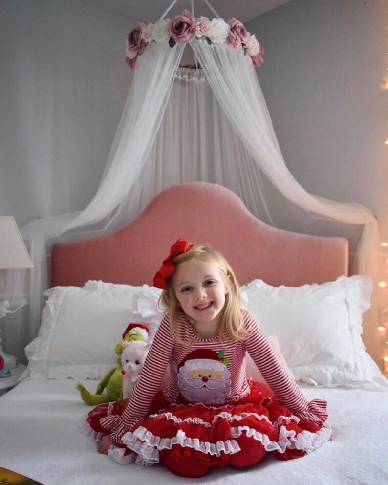 May include: A young girl wearing a red and white striped Christmas outfit with a Santa Claus graphic. She is sitting on a bed with a pink headboard, white pillows, and a floral canopy. A stuffed animal is next to her.