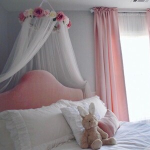 May include: A bedroom scene featuring a pink upholstered headboard, white bedding, and a plush bunny. A canopy with white fabric and a floral crown hangs above the bed. Pink curtains frame a window, creating a soft, inviting atmosphere.