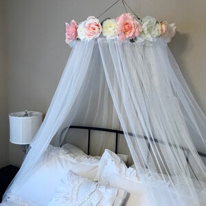 May include: A white canopy bed frame with a sheer, flowing white fabric draped over the bed. The canopy is adorned with a ring of large, colorful artificial flowers in shades of pink, white, and peach. The bed is made with white linens and pillows.