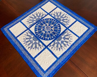 May include: A blue and white quilted table runner with a circular design in the center and four smaller squares around it. The runner is on a dark wood table.