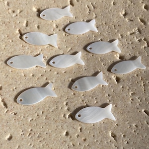 May include: A collection of nine small, white, iridescent fish-shaped charms made of mother-of-pearl. Each individual fish charm features a smooth, polished surface and a single small circular hole near the head for attachment or threading.