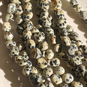 May include: A collection of multiple strands of dalmatian jasper beads. Each bead is irregularly shaped with a smooth, polished finish, featuring a cream-coloured base with distinct, scattered black and dark brown spots throughout the surface.