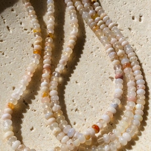 May include: A long strand of small, translucent, round beads in shades of pale cream, milky white, and soft pink, interspersed with occasional vibrant orange and amber-coloured beads, arranged in a multi-layered loop formation.
