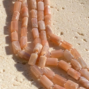 May include: A multi-strand necklace featuring cylindrical peach-coloured moonstone beads. The smooth, semi-translucent stones are strung together with small clear spacer beads, creating a layered appearance with a soft, reflective lustre.