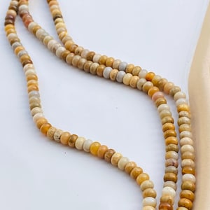 May include: A long strand of small, round, smooth beads in various shades of yellow, tan, beige, and light brown. The beads are strung together on a thin cord, displaying a natural, mottled appearance with subtle colour variations throughout the strand.