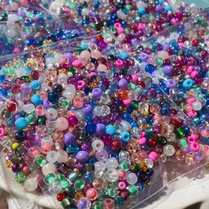 May include: A selection of multi-coloured beads in diverse forms, such as round, oval, and faceted. The beads feature hues of pink, purple, blue, green, and gold. They are packaged in clear plastic bags, suitable for jewellery making and crafts.