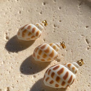 May include: Three small seashell pendants featuring a white base with brown spotted patterns. Each shell is fitted with a small gold-toned metal bail at the top, designed for attachment to a necklace chain or cord, showcasing natural textures.