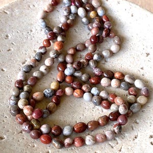 May include: A long strand of polished, oval-shaped beads in various earthy tones including deep red, brown, grey, and cream. The beads feature natural mottled patterns and are strung together, creating a continuous loop of smooth, multi-coloured stones.
