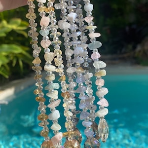 May include: Close-up of several strands of gemstone and crystal beaded jewellery. The strands feature a variety of colours, including yellow, pink, blue, and clear crystals. Each strand is adorned with a large, faceted crystal at the bottom.