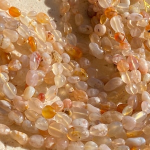 May include: A collection of smooth, tumbled gemstone beads in shades of translucent white, pale peach, and vibrant orange. The beads are irregularly shaped and polished, featuring natural inclusions and variations in colour, strung together on a thin yellow cord.