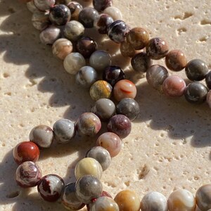 May include: A strand of round, polished beads featuring a variegated pattern of earthy tones including deep red, chocolate brown, cream, grey, and ochre. The beads are strung together, displaying natural swirling inclusions and varied colour gradients.