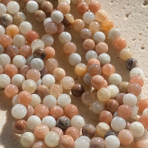 May include: A collection of round, polished moonstone beads in shades of peach, cream, and translucent white. The beads are strung together on a thin orange cord, displaying natural variations in colour, opacity, and subtle internal light reflections.