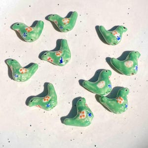 May include: A collection of ten small, light green ceramic bird-shaped beads. Each bead features a glossy finish with hand-painted orange floral patterns and small blue accents. The birds are arranged in a scattered pattern on a white surface.