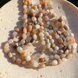 May include: A collection of smooth, polished, oval-shaped moonstone beads in shades of translucent white, cream, peach, and soft grey. The beads are strung together on thin orange thread, displaying a natural lustre and varied semi-opaque tones.