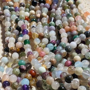 May include: A collection of multi-coloured round gemstone beads strung on thin threads. The beads feature a variety of translucent and opaque finishes in shades of green, purple, blue, pink, white, and brown, with some showing natural inclusions.
