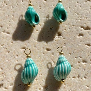 May include: Four small sea shell charms in a vibrant turquoise and white striped pattern. Each shell is accented with a small gold-toned metal jump ring at the top and a tiny gold-toned bead at the base, creating a set of decorative jewellery components.