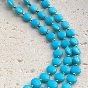 May include: A multi-strand necklace featuring smooth, oval-shaped turquoise beads in a vibrant blue hue, interspersed with small, round metallic gold-toned spacer beads. The necklace is arranged in a layered, curved formation.
