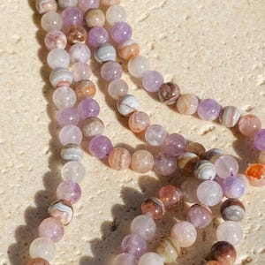 May include: A strand of round, polished beads in shades of lavender, soft purple, translucent white, and earthy brown. The beads feature natural banded patterns and are strung together on a thin purple cord, creating a multi-layered, circular arrangement.